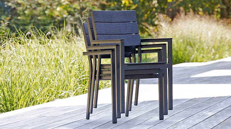 Drei schwarze stapelbare Gartenstühle mit Aluminiumgestell und breiten Lattenlehnen auf einer Holzterrasse vor hohem Ziergras.