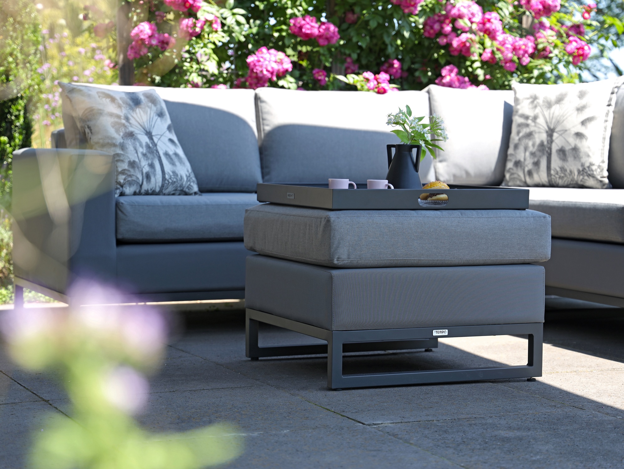 Moderne graue Lounge-Sitzgruppe mit Polsterhocker als Tisch auf einer Terrasse, umgeben von blühenden Rosen im Sommergarten.